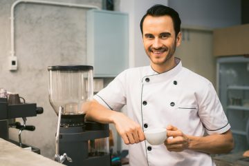 Indoor shot of young male barista making a cup of coffee while standing behind cafe counter. Young man pouring milk into a cup of coffee. Vintage filtered image.